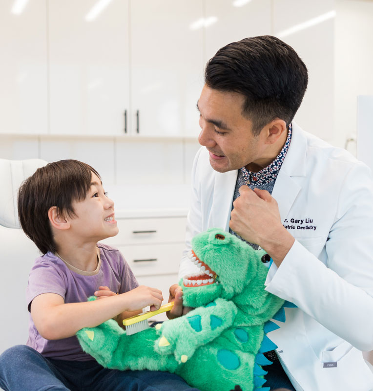 Child smiling while interacting with Dr. Gary Liu, holding a dinosaur toy and toothbrush in a pediatric dental office, promoting anxiety-free dental care for kids.