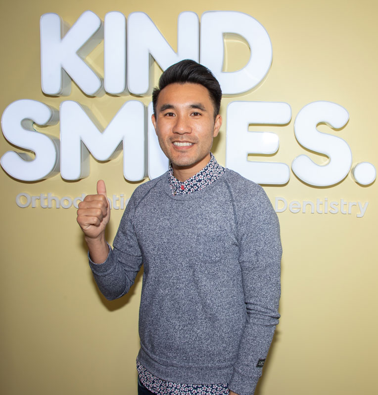 Smiling pediatric dentist giving a thumbs-up in front of the Kind Smiles logo, promoting affordable and compassionate dental care for children.
