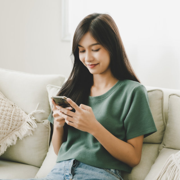 Young woman sitting on a couch, smiling while using a smartphone, representing convenience in contacting Kind Smiles dental practice.