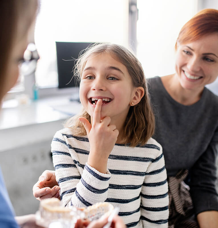 Young girl smiling and pointing to her teeth during a pediatric dental consultation, with a parent present, emphasizing the importance of dental health and restorative treatments for children.
