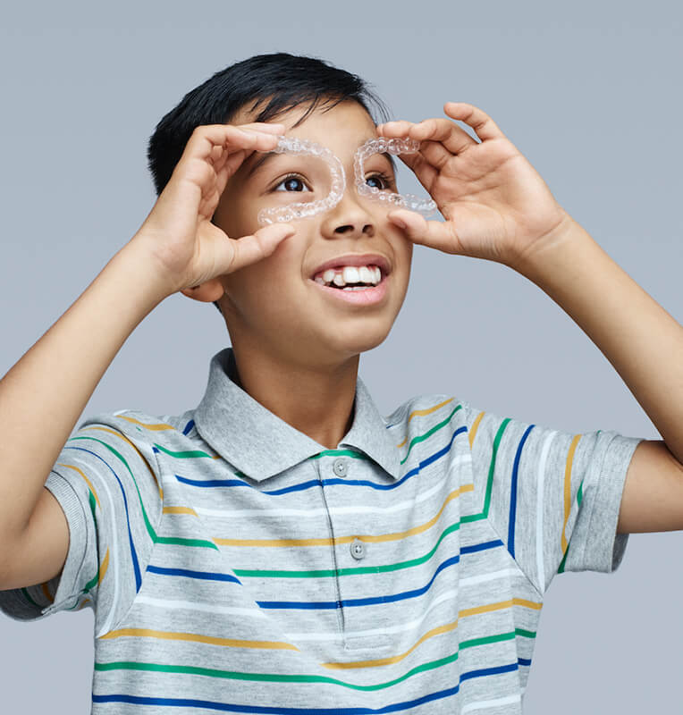 Boy playfully holding Invisalign® aligners like glasses, smiling, wearing a striped polo shirt, promoting Invisalign® treatment for kids.