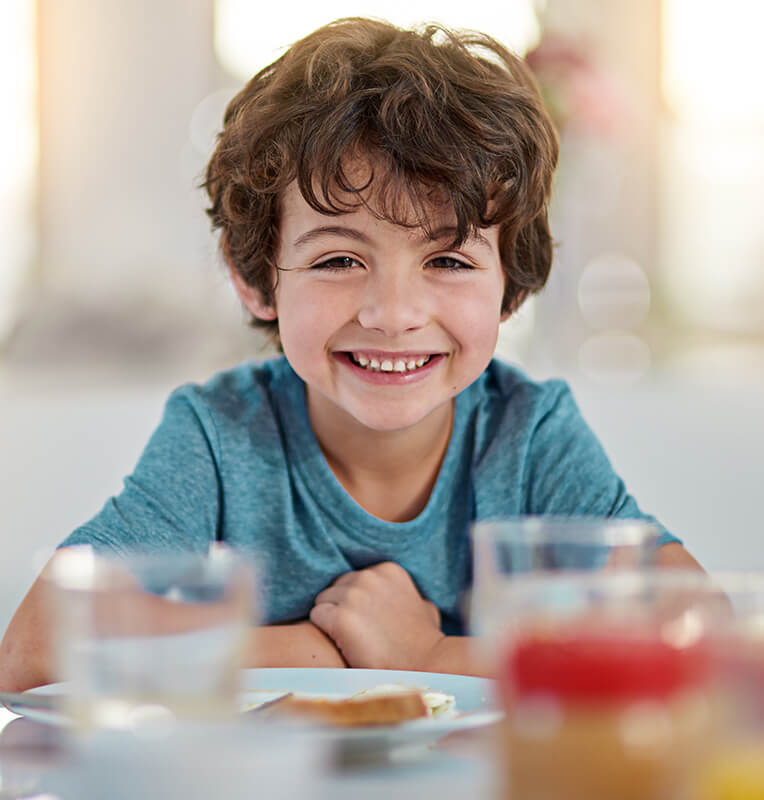 Smiling child in a blue shirt sitting at a table with a plate of food, representing the comfort and positivity associated with pediatric dental care at Kind Smiles Orthodontics.