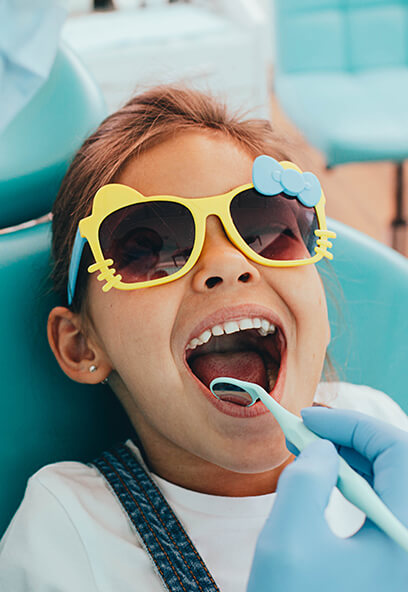 Child in colorful sunglasses smiling during a pediatric dental appointment, with a dentist using a dental tool, emphasizing a fun and friendly dental experience at KIND SMILES in Irvine, CA.