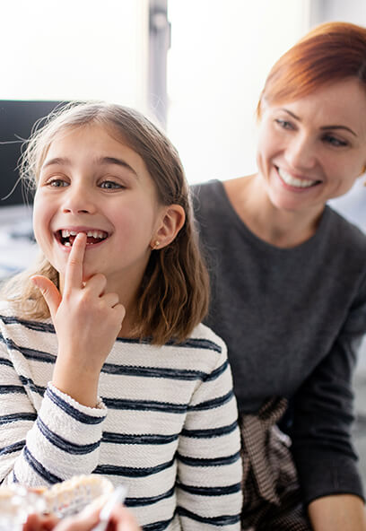 Smiling girl with a missing front tooth playfully pointing at her mouth, seated next to a smiling woman in a dental office, emphasizing a positive dental experience for children at KIND SMILES in Irvine, CA.