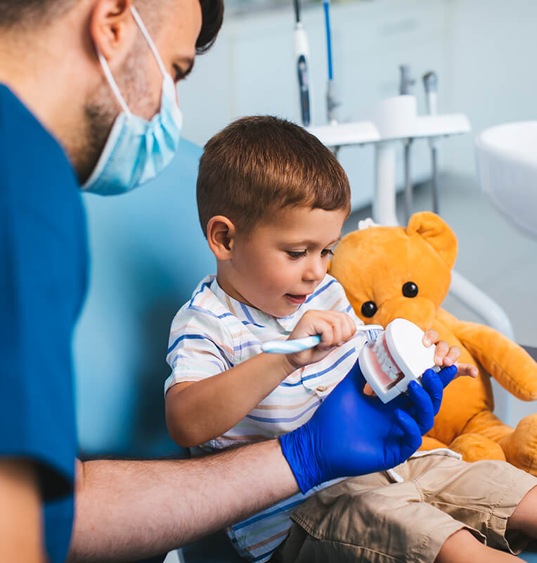 Child brushing a dental model with pediatric dentist in blue gloves, teddy bear nearby, promoting comfort during dental education at KIND SMILES Pediatric Dentistry.