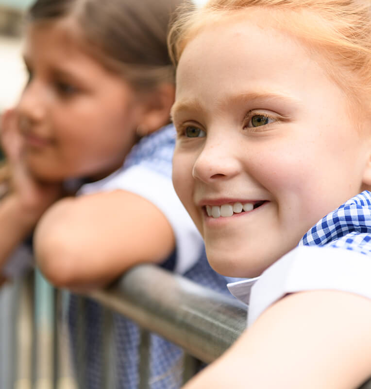 Smiling girl in a blue and white checkered shirt, enjoying a comfortable and friendly environment, with another girl in the background, reflecting the supportive atmosphere of special needs dentistry at Kind Smiles.